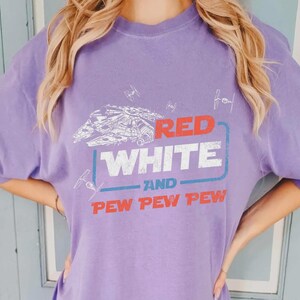 May include: A lilac-colored t-shirt featuring a graphic design with the text "RED WHITE AND PEW PEW PEW". The design includes a spaceship and starfighters. The shirt is a casual wear item.