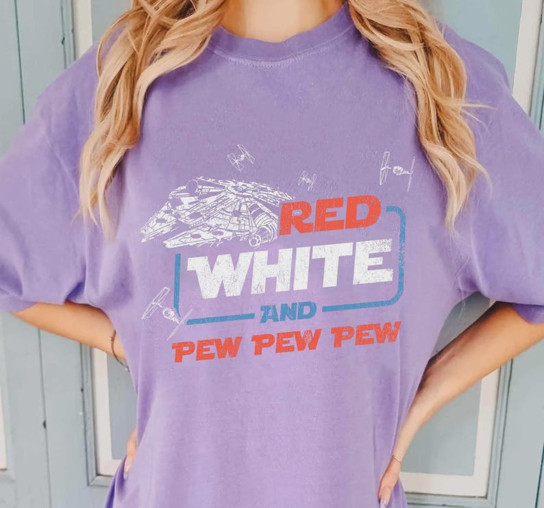 May include: A lilac-colored t-shirt featuring a graphic design with the text "RED WHITE AND PEW PEW PEW". The design includes a spaceship and starfighters. The shirt is a casual wear item.