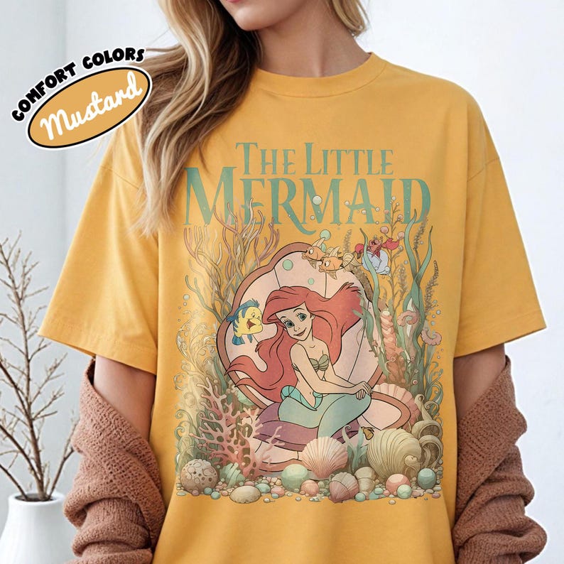May include: Mustard-colored t-shirt featuring a graphic of Ariel from The Little Mermaid. The design includes Ariel, Flounder, and Sebastian, set against an underwater scene with coral and seashells. The text "The Little Mermaid" is above the graphic.