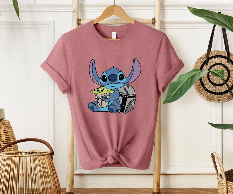 May include: A dusty rose t-shirt featuring a graphic of Stitch and Grogu from Star Wars. Stitch is blue with large ears, and Grogu is green. The design includes a silver helmet. The shirt has rolled sleeves and a knotted hem.