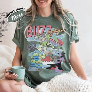 Puede incluir: Camiseta verde musgo con un gr&aacute;fico de Buzz Lightyear, el texto "Buzz Lightyear" y "Laser Blast". El dise&ntilde;o incluye extraterrestres y Zurg. La camiseta tambi&eacute;n tiene el texto "Comfort Colors".