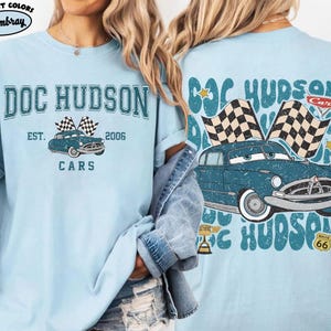 May include: Light blue t-shirt featuring a vintage-style graphic of a blue car with checkered flags, the text "DOC HUDSON", and "CARS". The shirt also includes the text "EST. 2006". The back of the shirt has a similar design.