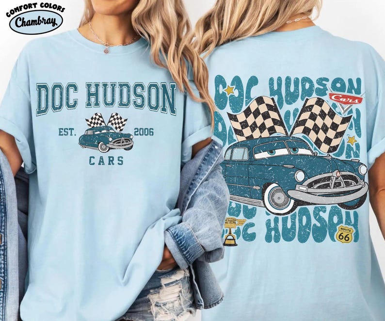 May include: Light blue t-shirt featuring a vintage-style graphic of a blue car with checkered flags, the text "DOC HUDSON", and "CARS". The shirt also includes the text "EST. 2006". The back of the shirt has a similar design.