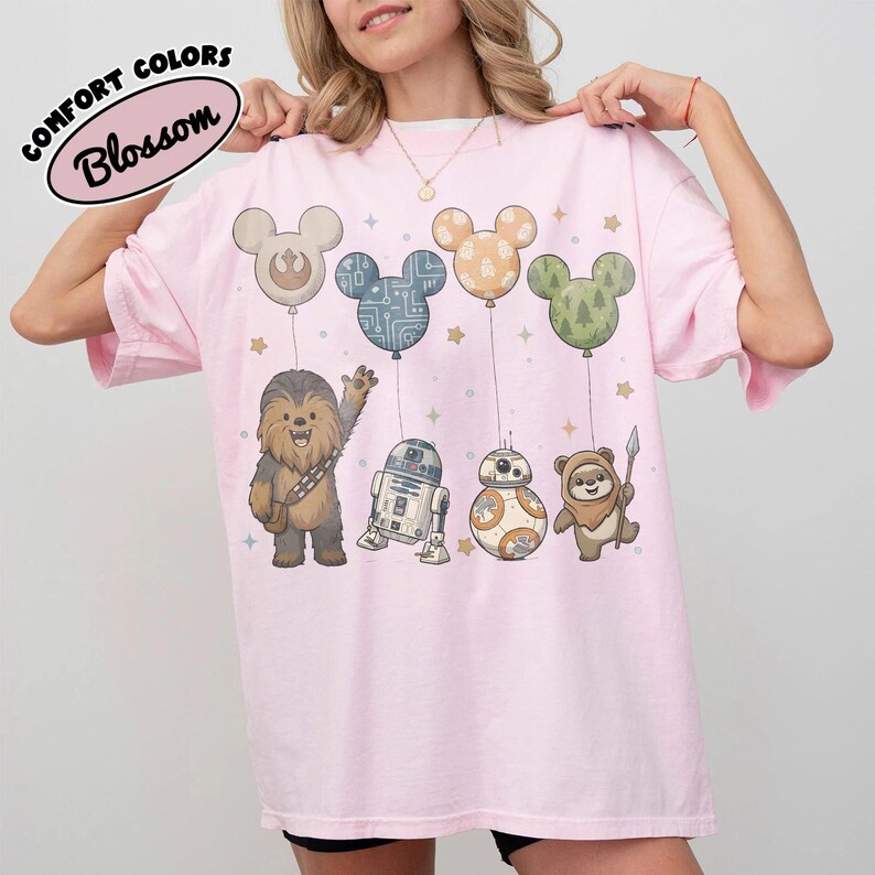 May include: A light pink t-shirt featuring cartoon characters from Star Wars, each attached to a Mickey Mouse-shaped balloon. The balloons are in various colors and patterns. The text "COMFORT COLORS Blossom" is in a white oval on the top left.