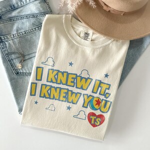 Puede incluir: Una camiseta de color crema con el texto azul y amarillo "I KNEW IT, I KNEW YOU" y un coraz&oacute;n rojo con las letras "TS". El dise&ntilde;o incluye peque&ntilde;as estrellas azules, contornos de nubes y un gr&aacute;fico de pelota que sustituye a la letra O.