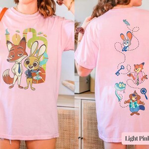 May include: Pale pink t-shirt with Zootopia cartoon characters. The front shows Nick Wilde and Judy Hopps. The back has various characters with a carrot, key, and magnifying glass. The text "Light Pink shirt" is at the bottom.