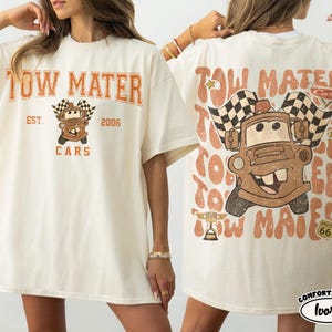 May include: Ivory-colored t-shirt featuring a distressed graphic of Tow Mater from the Cars movie. The front design includes the text "TOW MATER EST. 2006 CARS". The back design repeats "TOW MATER" with a cartoon image of Mater and checkered flags.