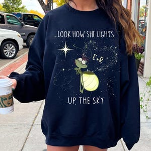 May include: Navy blue sweatshirt featuring a cartoon firefly holding a lantern, with the text "...LOOK HOW SHE LIGHTS UP THE SKY" and the initials "E+R". The design is set against a starry background.