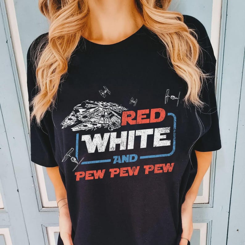 May include: Black t-shirt featuring a graphic design with the text "RED WHITE and PEW PEW PEW". The design includes a spaceship and fighter jets in red, white, and blue colors. The shirt is a crew neck style.