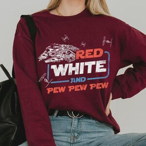 May include: Burgundy long-sleeve sweatshirt featuring a graphic design with the text "RED WHITE AND PEW PEW PEW". The design includes a spaceship and other space-themed illustrations in white and blue.