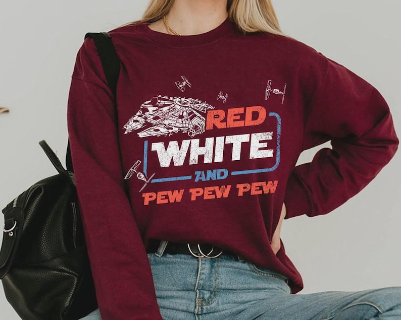 May include: Burgundy long-sleeve sweatshirt featuring a graphic design with the text "RED WHITE AND PEW PEW PEW". The design includes a spaceship and other space-themed illustrations in white and blue.