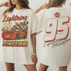 Op de afbeelding: Ivoor kleurig T-shirt met een Lightning McQueen-afbeelding uit de film Cars. De voorkant toont "Lightning 95 McQueen" met een geblokte vlag en een rode raceauto. De achterkant heeft een grote "95" en "Lightning McQueen".