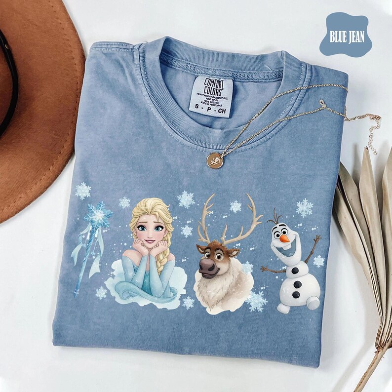 Peut inclure: T-shirt bleu jean avec Elsa, Sven et Olaf de La Reine des Neiges. Le t-shirt a un col rond et l'&eacute;tiquette de la marque Comfort Colors. Un collier en or avec un pendentif est drap&eacute; sur le t-shirt.