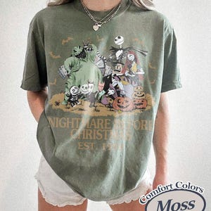 Puede incluir: Camiseta verde musgo con un dise&ntilde;o de "Pesadilla antes de Navidad". El dise&ntilde;o incluye personajes como Jack Skellington, Sally y Oogie Boogie, con calabazas y murci&eacute;lagos. El texto "NIGHTMARE BEFORE CHRISTMAS EST. 1993" est&aacute; impreso debajo.