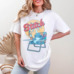 May include: White t-shirt featuring a cartoon illustration of Stitch from Lilo & Stitch relaxing on a beach chair with a drink. The word "Stitch" is written in a colorful, retro font above the character. The design is set against a yellow sun.