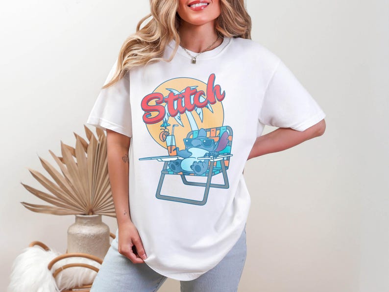 May include: White t-shirt featuring a cartoon illustration of Stitch from Lilo & Stitch relaxing on a beach chair with a drink. The word "Stitch" is written in a colorful, retro font above the character. The design is set against a yellow sun.
