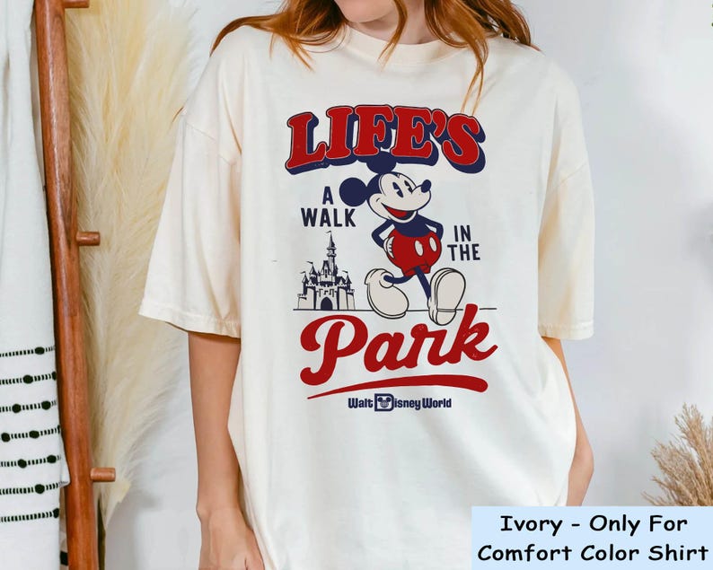 Puede incluir: Camiseta de color marfil con un gr&aacute;fico de Mickey Mouse y el texto "LIFE'S A WALK IN THE PARK" en rojo y azul. El dise&ntilde;o incluye un castillo y las palabras "Walt Disney World".