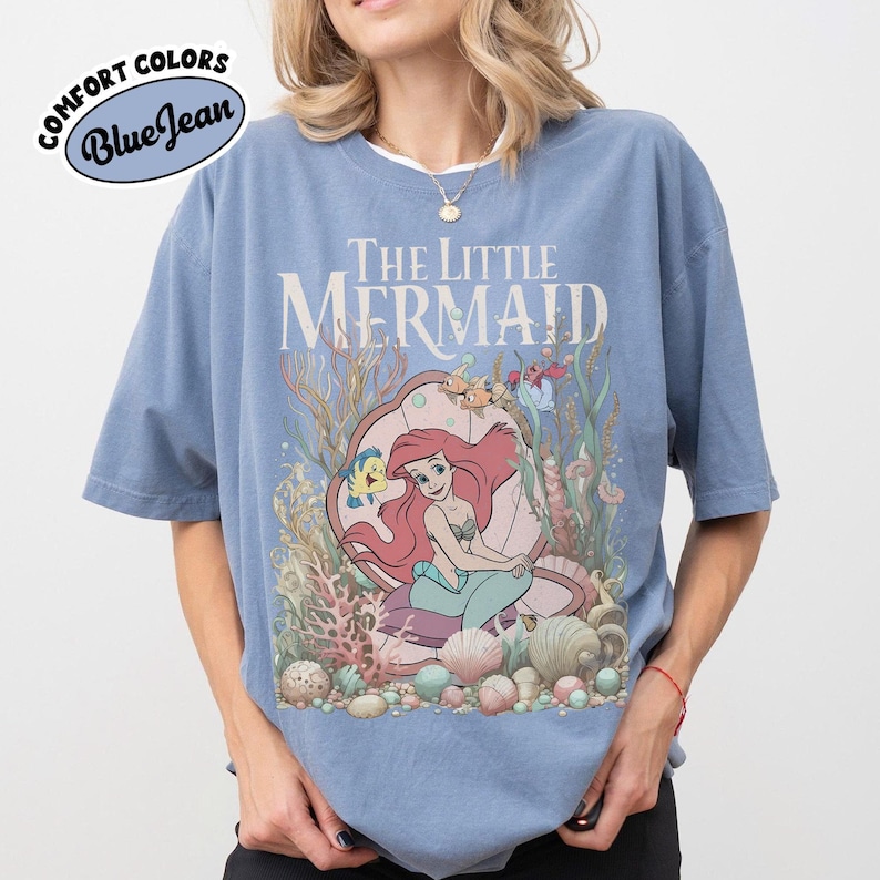 May include: A blue Comfort Colors t-shirt featuring a graphic from "The Little Mermaid." The design includes Ariel, Flounder, and Sebastian, surrounded by coral and seashells. The text "The Little Mermaid" is displayed above the graphic.