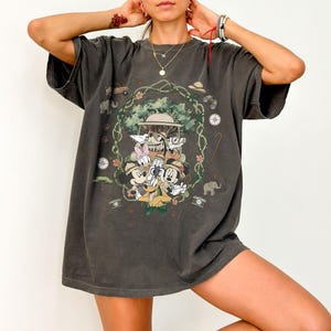 Puede incluir: Camiseta gris oscuro con un gr&aacute;fico de Mickey Mouse, Minnie Mouse, Donald Duck, Daisy Duck y Goofy con atuendos de safari. El dise&ntilde;o est&aacute; enmarcado por un borde floral verde y marr&oacute;n con iconos tem&aacute;ticos de safari. La camiseta tiene mangas cortas.