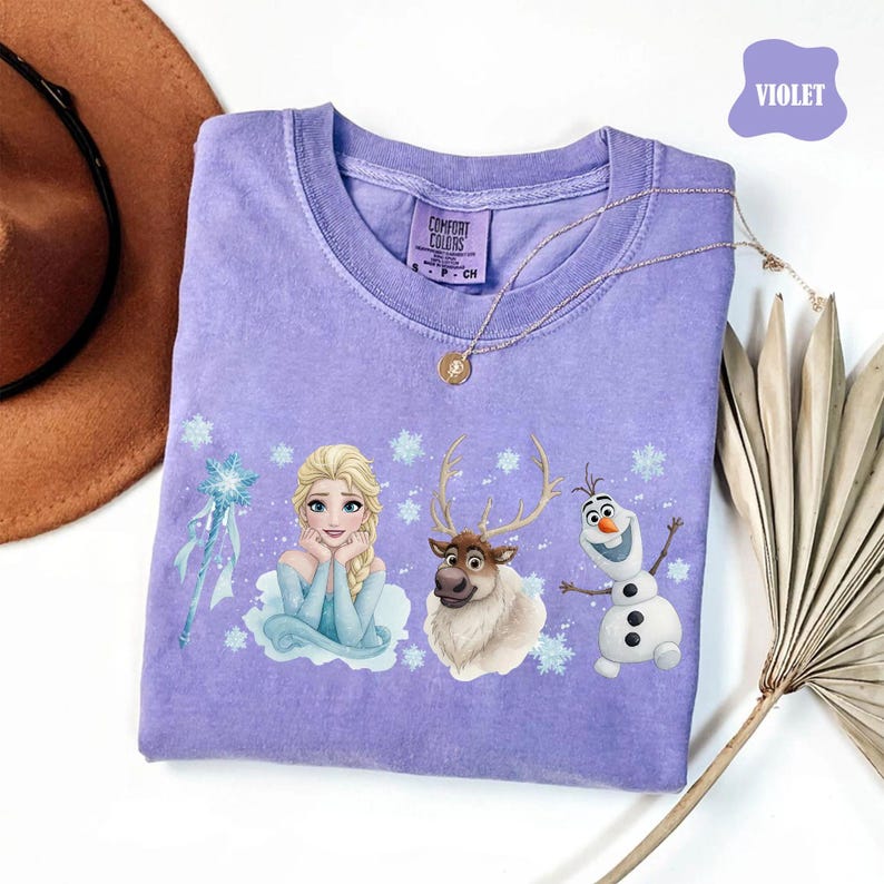 Peut inclure: Un t-shirt violet Comfort Colors avec un graphisme d'Elsa, Sven et Olaf du film La Reine des Neiges. Le motif comprend des flocons de neige et une baguette magique. Un collier en or est drap&eacute; sur le t-shirt.