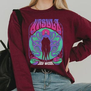 May include: Burgundy long-sleeve sweatshirt featuring a colorful graphic of the word "URSULA" above a stylized illustration of a sea witch with tentacles. The design includes shades of purple, green, and blue.