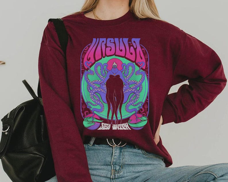May include: Burgundy long-sleeve sweatshirt featuring a colorful graphic of the word "URSULA" above a stylized illustration of a sea witch with tentacles. The design includes shades of purple, green, and blue.