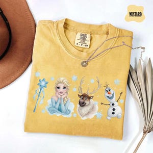 Peut inclure: T-shirt moutarde avec Elsa, Sven et Olaf de La Reine des Neiges. Le t-shirt a un col rond et des manches courtes. Un collier argent&eacute; est drap&eacute; sur le t-shirt. L'&eacute;tiquette indique "Comfort Colors".