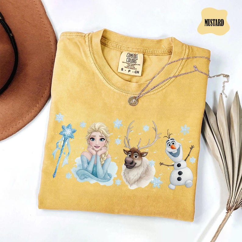 Peut inclure: T-shirt moutarde avec Elsa, Sven et Olaf de La Reine des Neiges. Le t-shirt a un col rond et des manches courtes. Un collier argent&eacute; est drap&eacute; sur le t-shirt. L'&eacute;tiquette indique "Comfort Colors".