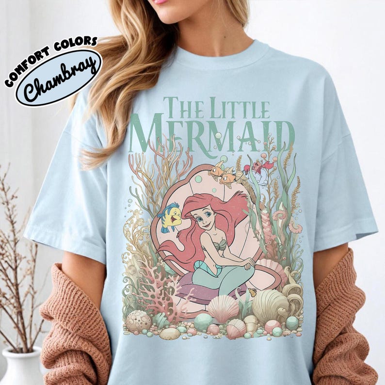 May include: Light blue t-shirt featuring a graphic of Ariel from The Little Mermaid. The design includes Ariel, Flounder, and other sea creatures within a seashell, with the words "The Little Mermaid" above. The shirt has the words "Comfort Colors Chambray" in a circle.