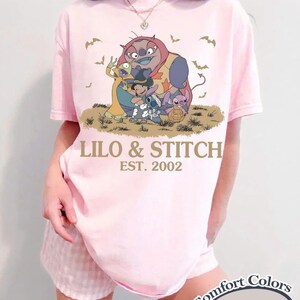 Op de afbeelding: Zachtroze T-shirt met een afbeelding van Lilo & Stitch-personages en de tekst "Lilo & Stitch EST. 2002." Het ontwerp bevat Stitch, Lilo en andere personages. Het shirt is een Comfort Colors Blossom.