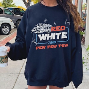 May include: Navy blue sweatshirt with a graphic design featuring a spaceship and the text "RED WHITE and PEW PEW PEW" in red and white. The design includes small starfighter illustrations. A casual, comfortable top.