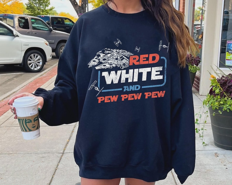 May include: Navy blue sweatshirt with a graphic design featuring a spaceship and the text "RED WHITE and PEW PEW PEW" in red and white. The design includes small starfighter illustrations. A casual, comfortable top.