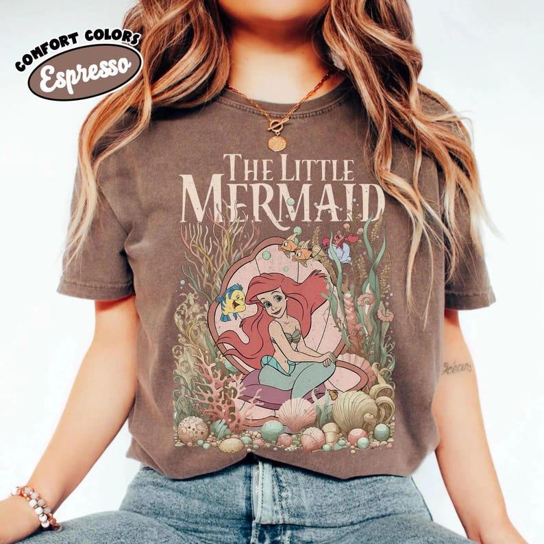 May include: Espresso-colored t-shirt featuring a graphic of Ariel from The Little Mermaid. The design includes the text "The Little Mermaid" above an illustration of Ariel in a seashell, surrounded by sea life and coral.