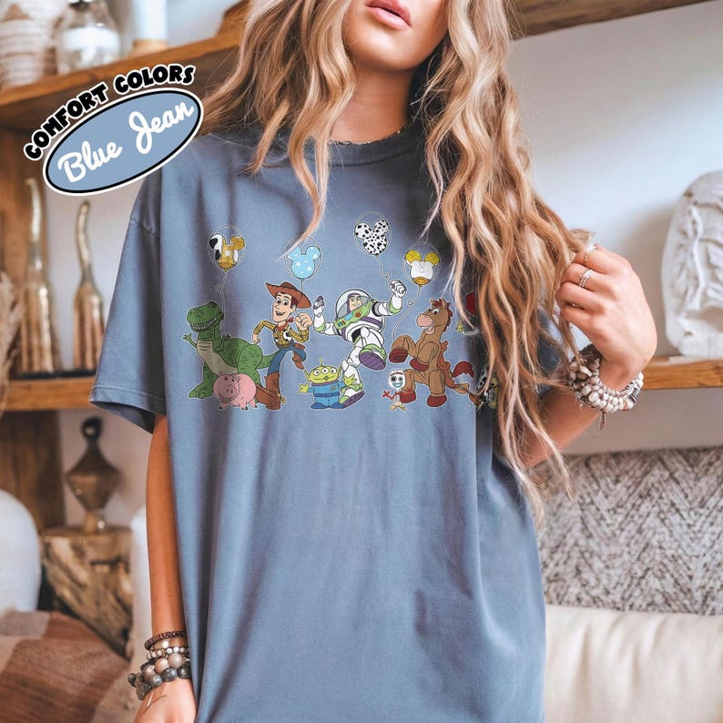 K&ouml;nnte beinhalten: Ein blaues T-Shirt mit einer Grafik von Figuren aus dem Animationsfilm "Toy Story". Das Design umfasst Woody, Buzz Lightyear, Rex und andere Charaktere. Das Shirt hat den Text "COMFORT COLORS Blue Jean" in einem kreisf&ouml;rmigen Design.