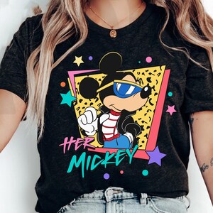 K&ouml;nnte beinhalten: Dunkelgraues T-Shirt mit einer Cartoon-Mickey-Mouse, die eine Sonnenbrille und eine schwarze Jacke tr&auml;gt. Die Grafik enth&auml;lt den Text "HER MICKEY" in einem farbenfrohen Retro-Stil mit Sternen und einem Leopardenmuster-Hintergrund.