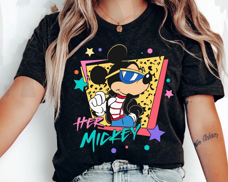 K&ouml;nnte beinhalten: Dunkelgraues T-Shirt mit einer Cartoon-Mickey-Mouse, die eine Sonnenbrille und eine schwarze Jacke tr&auml;gt. Die Grafik enth&auml;lt den Text "HER MICKEY" in einem farbenfrohen Retro-Stil mit Sternen und einem Leopardenmuster-Hintergrund.