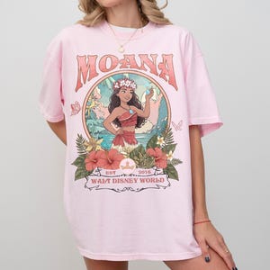 May include: Pink t-shirt featuring the word "MOANA" in a retro font above a circular graphic. The graphic depicts a cartoon character surrounded by flowers and the text "EST. 2016 WALT DISNEY WORLD".