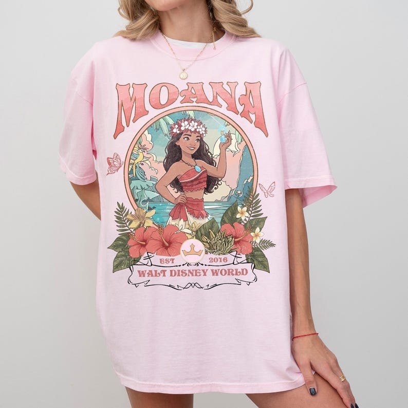 May include: Pink t-shirt featuring the word "MOANA" in a retro font above a circular graphic. The graphic depicts a cartoon character surrounded by flowers and the text "EST. 2016 WALT DISNEY WORLD".