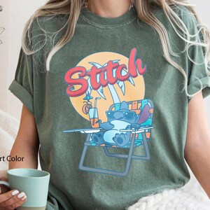 May include: Moss green t-shirt featuring a cartoon Stitch character relaxing on a beach chair, wearing sunglasses, and holding a drink. The word "Stitch" is written in red and orange above the character. The shirt is a comfort color.