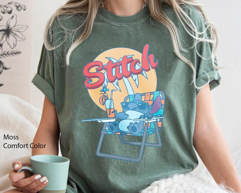 May include: Moss green t-shirt featuring a cartoon Stitch character relaxing on a beach chair, wearing sunglasses, and holding a drink. The word "Stitch" is written in red and orange above the character. The shirt is a comfort color.