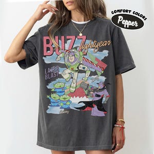 Puede incluir: Camiseta gris oscuro con un dise&ntilde;o de Buzz Lightyear de Toy Story. El dise&ntilde;o incluye el texto "Buzz Lightyear", "Laser Blast" y "Aliens", con un aspecto vintage y desgastado. La camiseta est&aacute; etiquetada como "Comfort Colors Pepper".