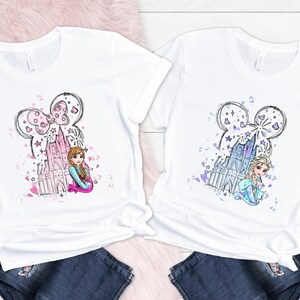 May include: Two white t-shirts with a cartoon graphic. One features a pink castle and a character with brown hair, the other a blue castle and a character with blonde hair. Both have a Mickey Mouse ear design.