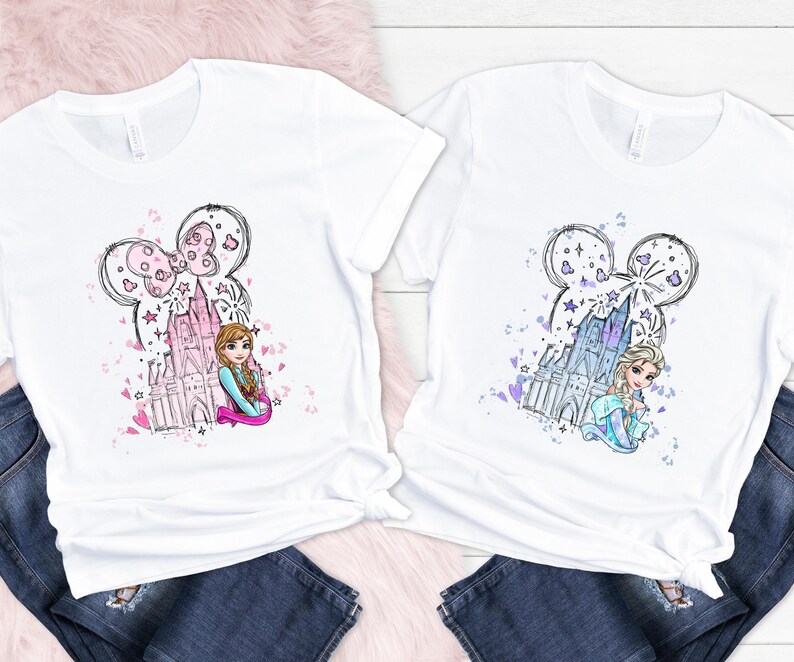 May include: Two white t-shirts with a cartoon graphic. One features a pink castle and a character with brown hair, the other a blue castle and a character with blonde hair. Both have a Mickey Mouse ear design.