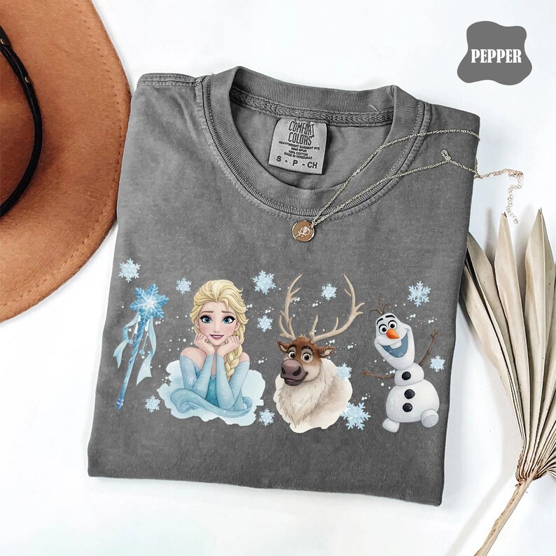 Peut inclure: Un t-shirt gris Comfort Colors avec Elsa, Sven et Olaf de La Reine des Neiges, avec des flocons de neige. Le t-shirt a un col rond et des manches courtes. Un collier argent&eacute; avec un pendentif est drap&eacute; sur le t-shirt.
