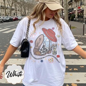 Peut inclure: Un t-shirt blanc &agrave; manches courtes avec une impression graphique repr&eacute;sentant une botte de cow-boy &agrave; motif vache, un chapeau de cow-boy rouge, des blocs empil&eacute;s avec les lettres T, S et un ciel bleu, et un collier de perles avec le texte I KNEW YOU WERE TROUBLE.