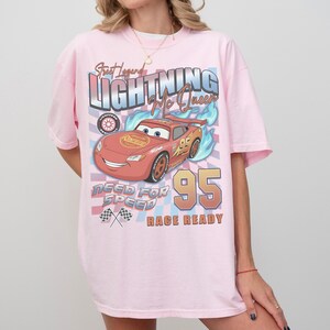May include: A light pink t-shirt featuring a graphic of Lightning McQueen from the Cars movie. The design includes the text "Street Legend Lightning McQueen", "Need for Speed", and "Race Ready", along with the number "95".