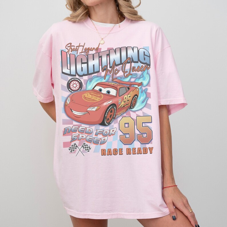 May include: A light pink t-shirt featuring a graphic of Lightning McQueen from the Cars movie. The design includes the text "Street Legend Lightning McQueen", "Need for Speed", and "Race Ready", along with the number "95".