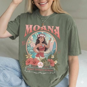 May include: Olive green t-shirt featuring a graphic of Moana from Walt Disney World. The design includes the word "MOANA" in pink, a circular illustration of Moana, and floral accents. The shirt has a relaxed fit.