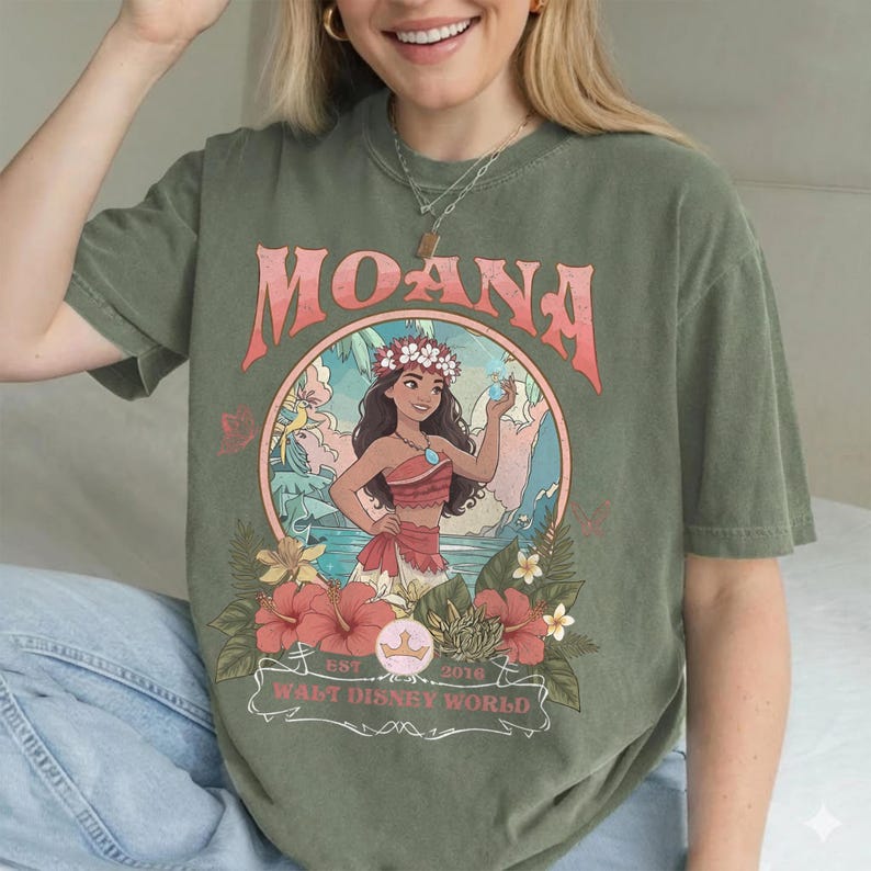 May include: Olive green t-shirt featuring a graphic of Moana from Walt Disney World. The design includes the word "MOANA" in pink, a circular illustration of Moana, and floral accents. The shirt has a relaxed fit.