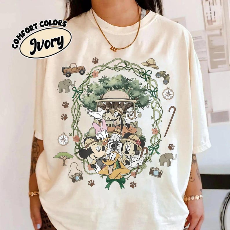 Puede incluir: Camiseta Comfort Colors color marfil con un dise&ntilde;o de tem&aacute;tica safari. El gr&aacute;fico presenta a Mickey Mouse, el Pato Donald y sus amigos, enmarcados por una corona verde con im&aacute;genes de safari como un jeep, una br&uacute;jula y unos prism&aacute;ticos. La camiseta tiene el texto "Comfort Colors" e "Ivory".